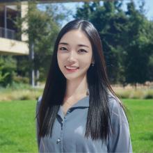 A professional headshot of Kelly Yoo, smiling outdoors with long straight black hair, wearing a light blue blouse. The background features a sunny green lawn and a building, creating a fresh and vibrant atmosphere.